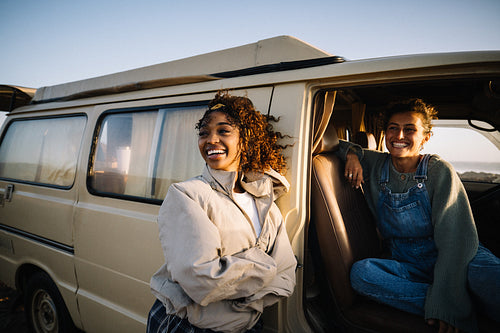 Two friends smile and laugh as they sit near a vintage van on a sunny day