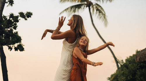 Woman and child dancing with arms waving in a tropical setting with trees at sunset
