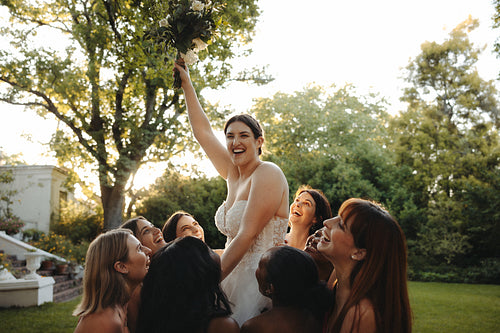 Joyful bride holding flowers surrounded by excited bridesmaids outdoors