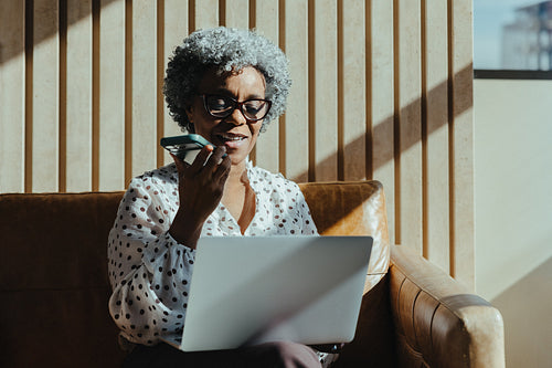 Mature professional woman multitasking with smartphone and laptop indoors