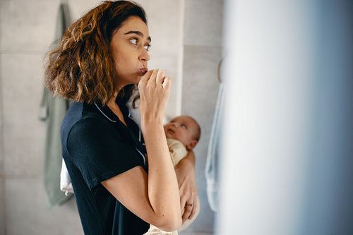 Mother brushes her teeth while holding a baby
