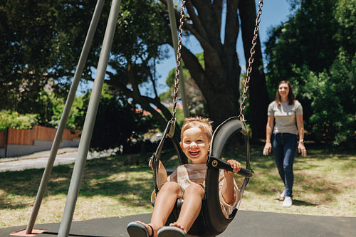 Child enjoying a swing in a sunny outdoor park with guardian nearby