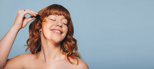 Happy woman with ginger hair applying face serum using a dropper in a studio