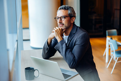 Thoughtful businessman sitting at desk by window with laptop and mug
