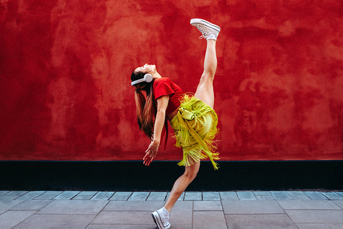 Young woman kicks into dance with headphones beside a red wall