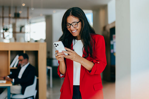 Smiling woman in red blazer checking her phone in office environment
