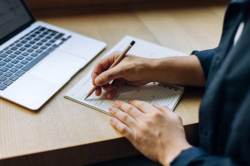 Person writing in a notebook with a laptop on a wooden desk