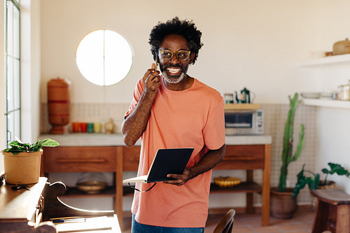 Afro-Brazilian man in 50s working from home, holding mobile phone and notebook in kitchen