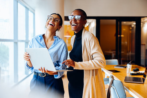 Two women laughing and using a laptop in a bright office setting