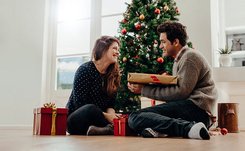Young couple exchanging Christmas gifts sitting beside a Christmas tree at home. Man giving a Christmas present to his girlfriend.