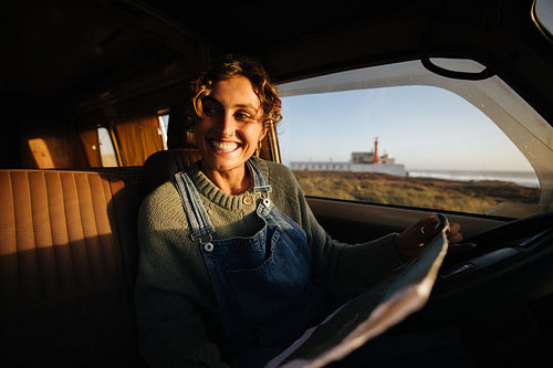 Smiling woman drives car with map near coast and lighthouse