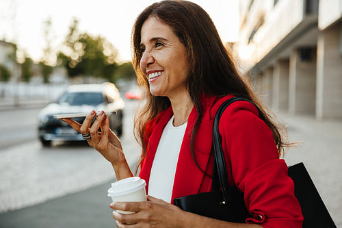 Middle aged woman using voice commands on phone while holding coffee in city