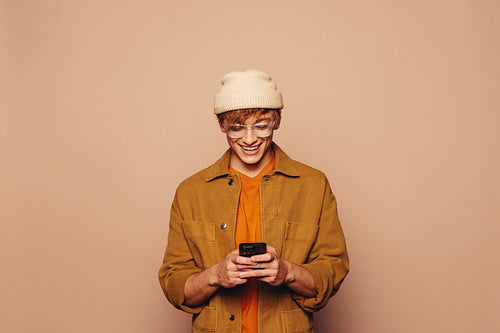 Cheerful young man smiling while reading messages on a smartphone against a vibrant peach background