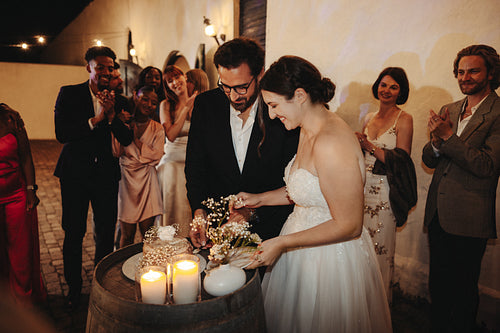 Smiling newlyweds cutting cake surrounded by joyful guests during their wedding celebration