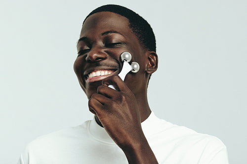 Happy black man massaging his face with a 3D roller as part of his skincare routine