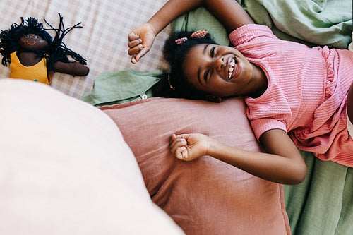 Happy black girl playfully relaxing on bed with her doll nearby