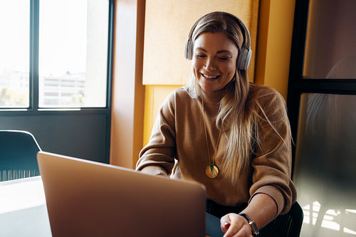 Smiling young woman working on a laptop while wearing headphones indoors