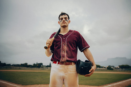 Confident baseball player standing on the field holding a bat and helmet, ready for the game