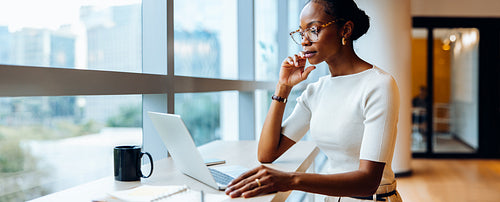Woman using laptop and thinking while working near large window