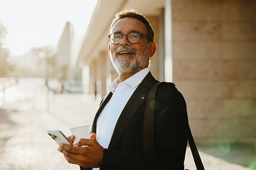 Senior businessman outdoors holding smartphone and coffee cup with a cheerful mood