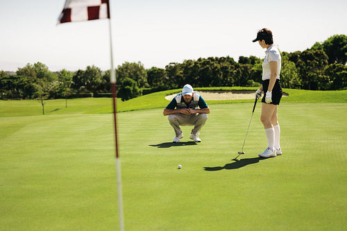 Woman golfer aiming on putting green near flagstick as male player observes
