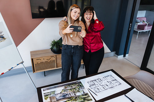 Two women taking a selfie in an office with architectural plans