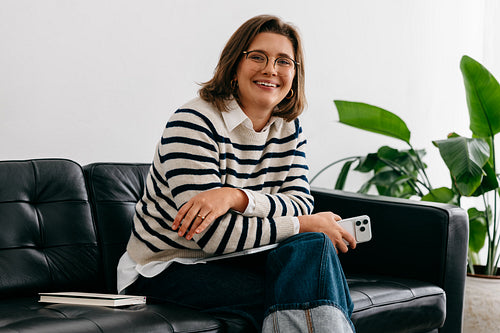 Woman smiling at camera with smartphone in relaxed home setting