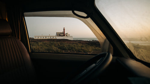 A quiet roadside drive frames a distant lighthouse on the coast through a car window at sunset