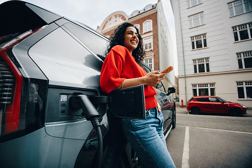 Waiting beside an electric car, a woman uses her smartphone in the city