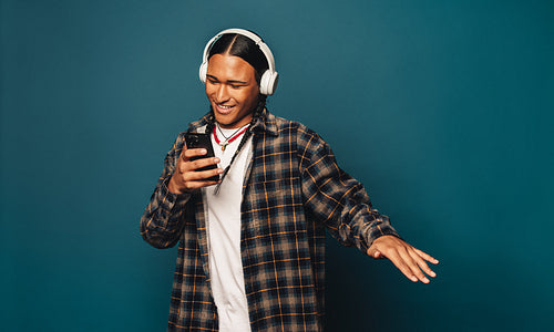 Happy young man dancing to music on blue background with braided hair and smartphone