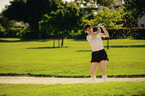 Female pro golfer in action hitting the ball from a sand bunker in the glow of the sun