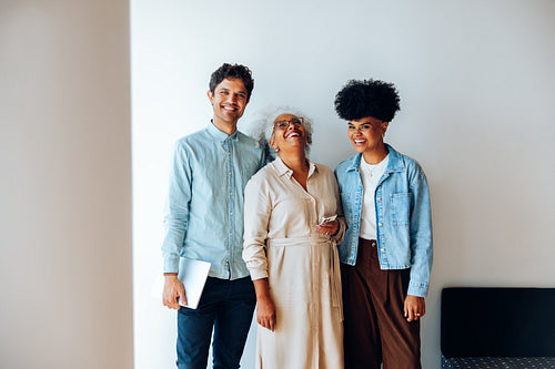 Three professionals laugh together in office hallway with laptop and phone