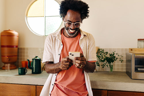 Brazilian man with afro hair smiling and texting on a mobile phone in the kitchen