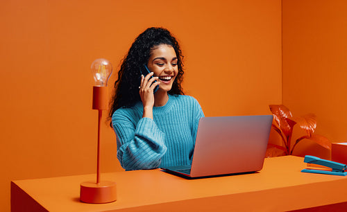 Happy female entrepreneur with curly hair speaking on a mobile phone, working on laptop in a creative, minimalistic, color-blocking workspace