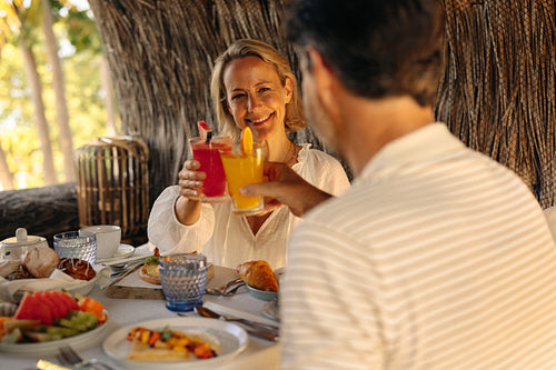 Happy couple enjoying luxurious breakfast at a resort, toasting with drinks under a shaded area