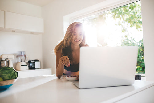 Woman in kitchen eating berries and using laptop