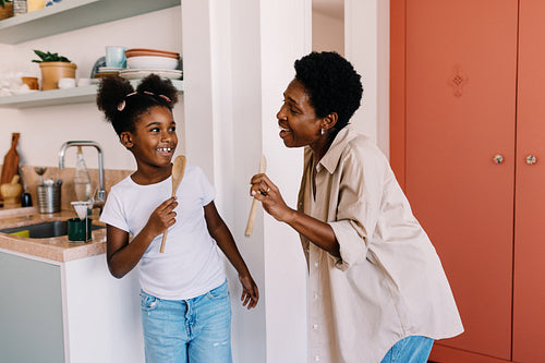 Happy mother and daughter singing with wooden spoons in the kitchen