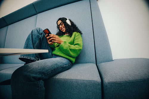 Young woman relaxing with smartphone in study lounge wearing headphones