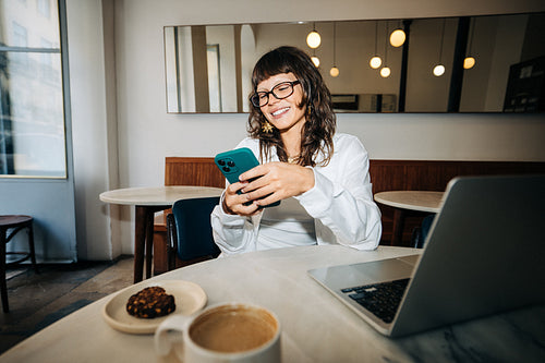 Smiling woman using smartphone in cafe with laptop
