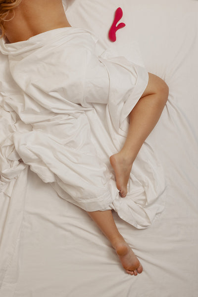 Woman lying on a bed with white covers, exploring erotic pleasure with a vibrator