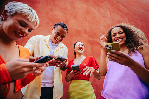Friends swap mobile screens and laugh together outdoors by a colorful wall