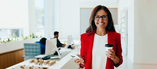 Confident smiling woman holding coffee and phone in a bright office setting