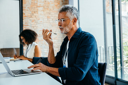 Mature businessman speaking on phone while using laptop in modern office setting with colleague working in background