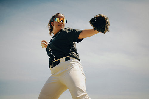 Confident female baseball pitcher about to throw the ball wearing sunglasses and a black jersey