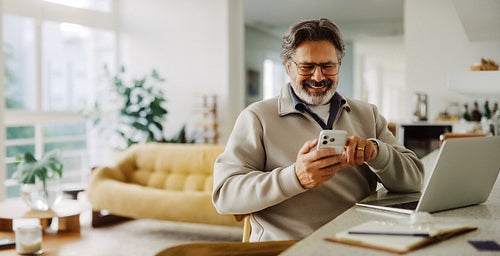 Senior man uses smartphone at home with a laptop