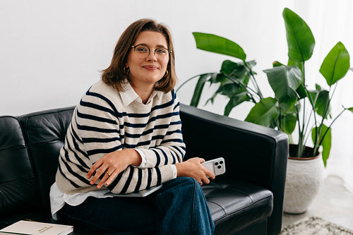 Woman smiling at camera while using smartphone on couch during remote work