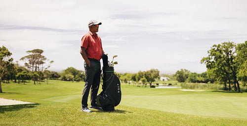 Professional golfer standing on a golf course, holding a bag of golf clubs in wide shot of golf resort