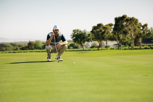 Crouching golfer on putting green aiming at golf ball on a lush green course under a clear sky, balancing on a club