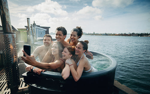 An outdoor scene shows friends taking a selfie with a phone in a hot tub on vacation.