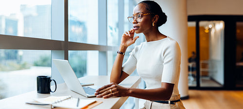 Professional woman working on her laptop while standing near large office windows